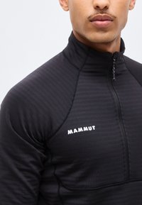 Mannequin masculin portant un haut de sport Mammut noir à demi-fermeture éclair, montrant la partie supérieure de la poitrine et du cou avec la fermeture éclair fermée.