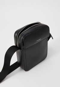 Bolso cruzado de cuero negro con cierre de cremallera, bolsillo frontal plano y correa de tela ajustable; diseño minimalista y textura suave.