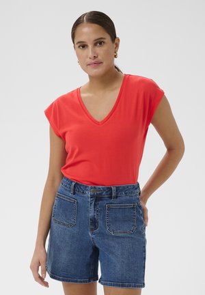 Femme aux cheveux bruns attachés en queue de cheval, portant des boucles d'oreilles créoles dorées, un t-shirt rouge à col en V et un short en jean bleu avec des poches avant.