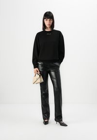 Sweatshirt preto de malha com pequenos adornos, combinado com calças de couro pretas. A modelo segura uma mala creme com detalhes de logotipo e usa sapatos de salto agudo.