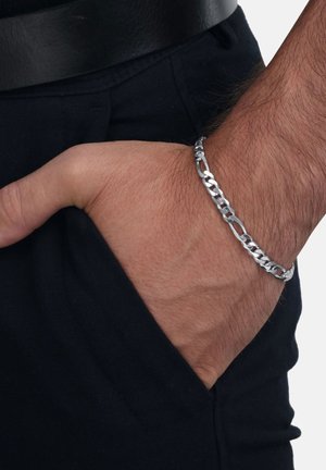 Mano masculina con una pulsera de cadena plateada descansando dentro del bolsillo frontal de unos pantalones negros, contra un fondo blanco.