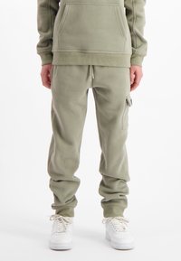 Persoon in olijfgroene joggerbroeken met zijzak en witte sneakers, staand tegen een witte achtergrond.