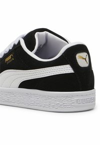 Crne suede tenisice s bijelim detaljima; imaju teksturiranu gumenu potplat i zlatni Puma natpis na peti i bočnoj strani.