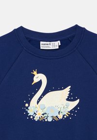 Sweatshirt azul marinho com um cisne branco a usar uma coroa dourada rodeado por flores azul claro e amarelas impressas à frente.