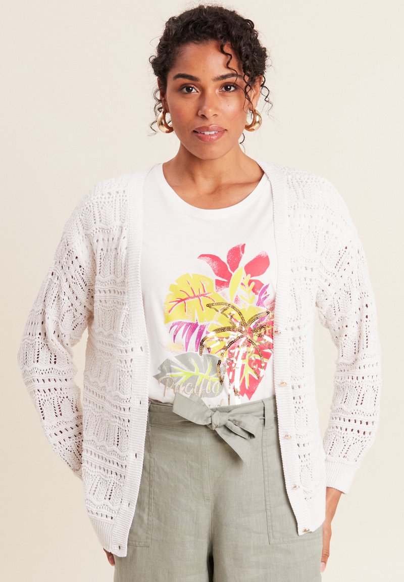Weiße Strickjacke mit Lochmuster, getragen über einem weißen T-Shirt mit buntem tropischem Blattdruck und grauen hochgeschnittenen Hosen mit Schleife.