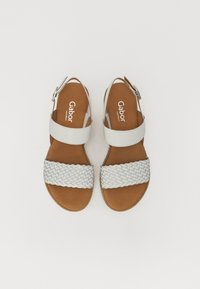 White sandals featuring a woven strap design, smooth upper band, and a brown padded insole. Adjustable ankle strap with silver hardware.