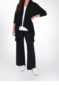 Cardigan leggero nero con frontale drappeggiato aperto, abbinato a una maglietta bianca a V e pantaloni neri a gamba larga, completato da sneakers bianche.