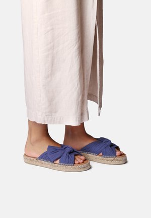 Fødder iført blå stof-slip-onsandaler med sløjfedetalje og vævede såler, kombineret med beige bukser med vide ben og sideslids.