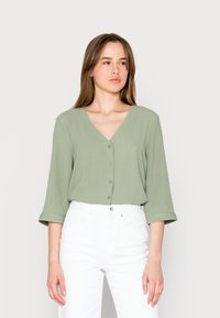 Lys grønn V-halsbluse med trekvart ermer og frontknapper, kombinert med hvite høytlivsjeans. Glatt tekstur, uformelt design.