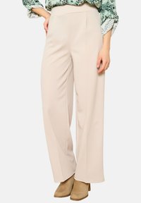 Béžové široké kalhoty z hladkého materiálu s vysokým pasem, s jemnými předními sklady a přizpůsobeným střihem, nošené s hnědými botami na podpatku.