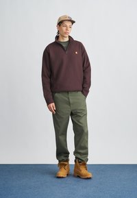 Pull-over en polaire bordeaux avec une demi-fermeture éclair, pantalon vert foncé, et bottes marron clair avec des lacets jaunes, sur un fond gris clair.
