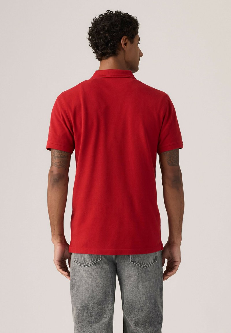Homme aux cheveux bouclés portant un polo rouge uni à manches courtes et un jean gris clair, debout dos au spectateur.