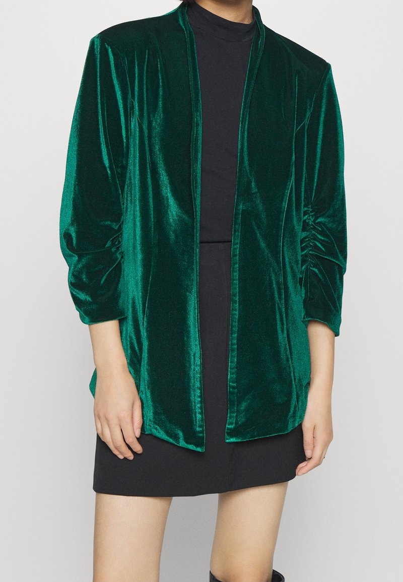Femme portant une veste en velours vert émeraude brillant à ouverture devant avec des manches froncées, par-dessus une robe noire, sur un fond uni.