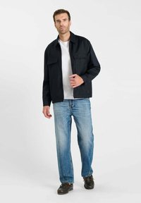 Donkerblauwe jas met knopen en twee borstzakken, gedragen over een wit T-shirt en lichtblauwe spijkerbroek. Model staat met een neutrale uitdrukking.