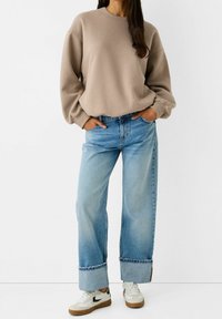 Mujer vestida con un sudadera beige de gran tamaño, vaqueros de pierna ancha azul claro con puños y zapatillas blancas con detalles en negro y marrón, con las manos en los bolsillos.