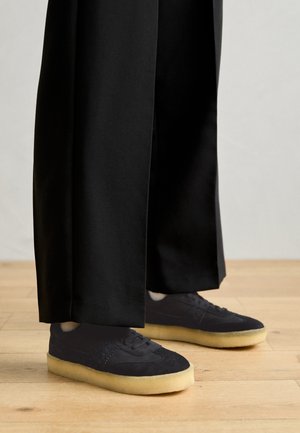 Schwarze Wildledersneaker mit einem strukturierten Design und einer cremefarbenen Gummisohle, kombiniert mit locker sitzenden schwarzen Hosen, stehend auf einem Holzboden.