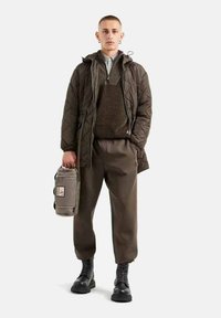 Giubbotto imbottito marrone sopra un maglione a maglia, abbinato a pantaloni larghi e stivali neri. Tiene una borsa grigia con un logo. Sfondo semplice.