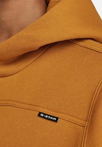 Sudadera color mostaza con una tela texturada, que cuenta con una gran capucha y una pequeña etiqueta negra de G-Star en la zona del pecho.