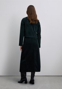 Tenue en velours côtelé vert foncé comprenant un haut court et une longue jupe. La jupe comporte une fente latérale, et le mannequin porte des bottines à talons noires. Texture lisse.