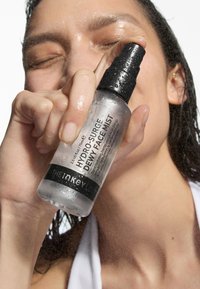 Hydro-Surge Dewy Face Mist in einer klaren Flasche mit schwarzem Sprühkopf. Wassertropfen auf der Flasche; mit einem Etikett, das den Produktnamen und die Details enthält.
