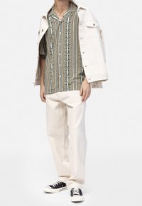 Short-sleeve patterned shirt in green, orange, and white, paired with cream pants and a white denim jacket. Black sneakers complete the look.