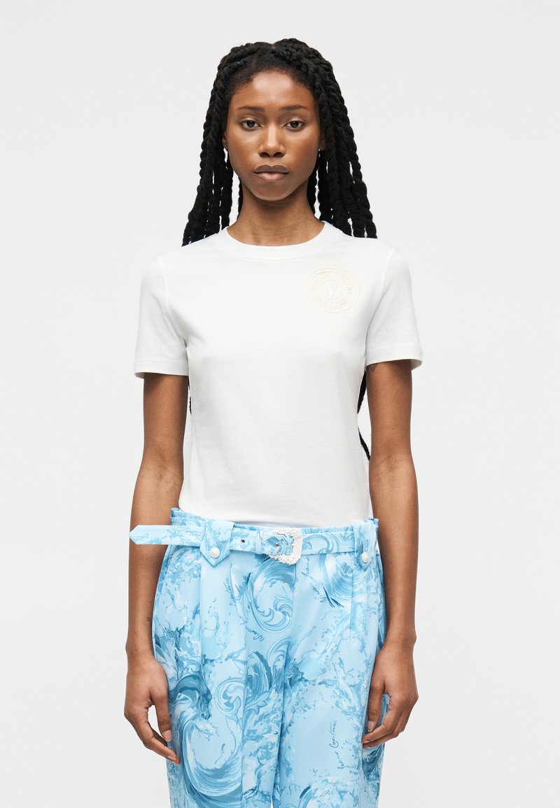 Γυναίκα με μακριά πλεξούδες, φορώντας λευκό πουκάμισο με κοντά μανίκια και μπλε παντελόνι με σχέδια και ζώνη που ταιριάζει, στέκεται μπροστά σε απλό φόντο.