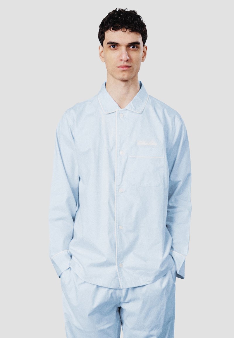 Jeune homme aux cheveux bouclés et foncés, portant un pyjama bleu clair boutonné à manches longues, les mains dans les poches, sur un fond uni.