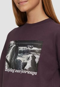 Paarse sweatshirt met een grafische print van een vrouw met lang haar die met een hond rijdt, met de tekst "Geniet van nieuwe avonturen" onderaan.