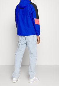 Modrá lehká bunda s černými a růžovými akcenty, nastavitelnou kapucí a elastickými manžetami. Nositelná přes světle modré džíny a bílé tenisky.