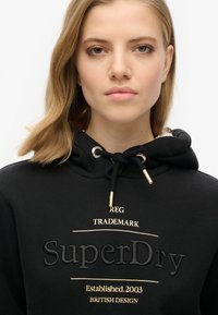 Sudadera negra con detalles dorados y logo bordado de "SuperDry". Cuenta con cordón ajustable y una textura de tela suave.