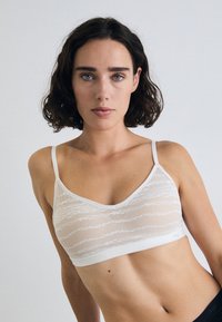 White bralette featuring a textured sheer fabric with wavy patterns, thin adjustable straps, and a wide elastic band under the bust.