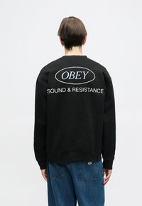 Črna majica s kapuco, ki prikazuje velik bel ovalni logotip z napisom "OBEY" in besedilom "SOUND & RESISTANCE" v belem tisku na zadnji strani.