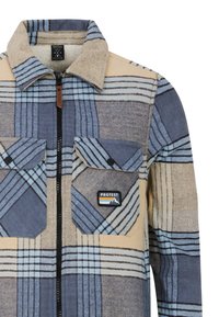 Chemise en flanelle à carreaux dans des tons de bleu et de beige, avec une fermeture zip à l'avant, deux poches poitrine et un patch logo. Texture douce et motif à carreaux.