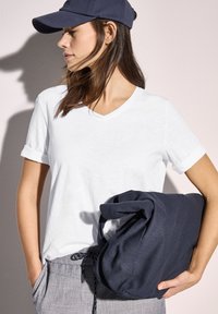 Witte v-hals t-shirt, marineblauwe pet en grijze met patroon broek. Houdt een marineblauwe stoffen tas vast met een zachte textuur en opgerolde handgrepen.