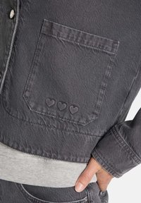 Graue Jeansjacke mit einer Brusttasche, die drei geprägte Herzformen aufweist, und grauen Rippbündchen. Glatte Textur, modernes Design.