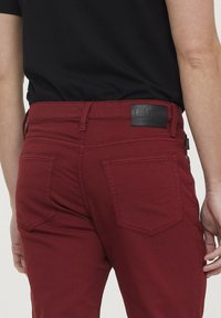 Pantalons en denim rouge avec une finition texturée, dotés de deux poches arrière, d'une étiquette de ceinture en cuir noir avec logo, et de passants de ceinture.