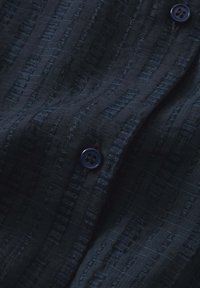 Tessuto navy a trama con strisce verticali e motivi sottili; presenta un bottone blu scuro, mettendo in evidenza un aspetto leggero.