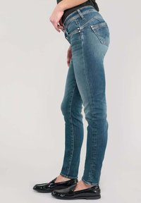 Jean en denim bleu marine, coupe slim, avec taille haute, cinq poches, un léger effet délavé et des détails de coutures. Associés à des chaussures noires.