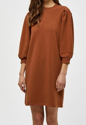 Femme portant une robe marron rouille longueur genoux avec des manches trois-quarts bouffantes et des poignets côtelés, debout devant un fond uni.