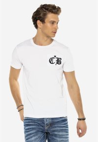 Weiße Baumwoll-T-Shirt mit einem schwarzen Textlogo auf einer Brusttasche. Kurzärmlig und mit Rundhalsausschnitt. Trägt man mit blauen Denim-Jeans.