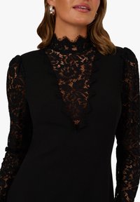 Černé šaty s hlubokým výstřihem do V z krajky a dlouhými krajkovými rukávy. Mají texturované krajkové detaily a padnoucí siluetu.