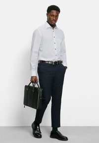 Witte shirt met kleine blauwe stippen, lange mouwen, zwarte knopen, gecombineerd met marineblauwe broek en gepolijste zwarte dresschoenen. Zwarte draagtas.
