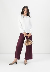 Bílá košile s dlouhými rukávy a texturovanými manžetami, tmavě fialové skládací široké kalhoty, šedé slingback podpatky a vícebarevná kabelka.
