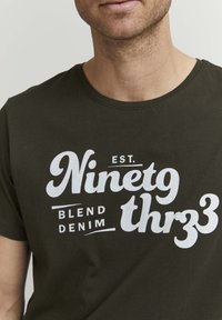Oliven grønn bomulls T-skjorte med stor hvit tekst som sier "ETABLERT. Nitti-Tre Blend Denim" i ulike skrifttyper og stiler.