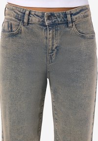 Augsta vidukļa izbalējušas zilas džinsa bikses ar jostas cilpām, pogas aizdari, priekšējām kabatām un redzamu baltu īsu topu virs jostasvietas.