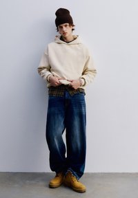 Beige hoodie över en brun rutig skjorta, i kombination med lösa blå jeans och gula stövlar. Bakgrunden är enkel och ljus.