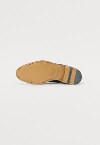 Suela de zapato de cuero en color marrón claro, acabada con detalles en negro, costuras visibles y detalles de marca. Superficie texturizada y forma redondeada.