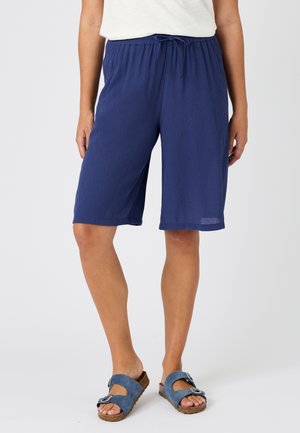 Persona che indossa pantaloncini larghi blu navy fino al ginocchio con vita a coulisse e sandali blu con fibbia su sfondo neutro.