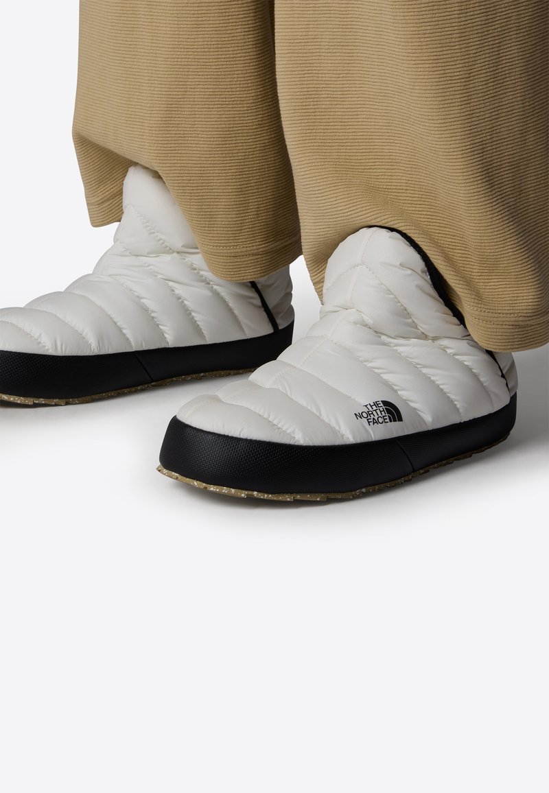 Bílé nadýchané bačkory s černými gumovými podrážkami a texturovaným gripem. Zahrnují prošívaný design a logové detaily na boku.