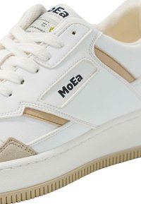 White sneaker with beige accents and suede detailing. Features a textured rubber sole and logo on the side. Fabric laces and tongue.